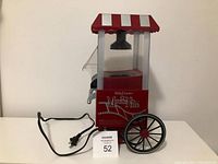 Front-side view of the red and white Betty Crocker Movie Nite popcorn maker, showing power cord, wheels, and clear panels