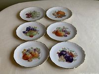 Six dessert plates laid out showing various fruit paintings and gold trim edges