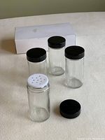 Eight clear cylindrical glass spice shakers with black lids, one white lid shaker with holes, arranged on a surface with the box in the background.
