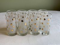 Eight clear drinking glasses decorated with gold polka dots and fine gold swirls arranged in two rows, viewed from the side.
