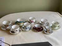 View of all nine teacups with their matching saucers arranged in two rows showing assortment of floral patterns and gold rims.