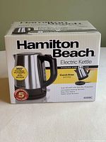 Photo of the closed box showing Hamilton Beach Electric Kettle branding, capacity 1.0 litre, stainless steel body, and features like 360° cordless portability and auto shutoff.