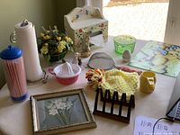 Overall view of all kitchen and decor items on table including glass cutting board, measuring cups, teapot cozy, strainer, egg timer, green bowl, paper towel and tissue holders, faux floral basket, straws dispenser, bud vase, figurines, juicer, framed print, multi-purpose holder.