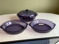 Three Pyrex purple glass pieces: covered serving dish and two pie plates displayed on white table.