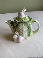 Photo showing side view of ceramic teapot shaped like lettuce with a white rabbit figurine on the lid and another rabbit near the spout base.