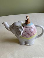Elephant-shaped teapot with monkey figurine lid, side view showing elephant trunk spout and decorative blanket pattern.