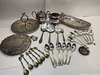 View showing assortment of silver plated serving trays, teapot, creamer, sugar bowl and various spoons and forks.