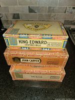 Stacked view of three vintage wooden cigar boxes with branded labels King Edward, John Carver, and Dukes on a tiled kitchen surface.