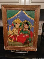 Full view of the framed Persian reverse glass painting depicting a seated couple in traditional dress against a blue background with green and gold curtain details.