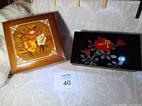 Two music boxes, one wooden with floral inlay and one lacquered with red floral decoration, placed side by side.