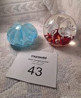 Two glass paperweights on fabric background with auction tag. One is blue with bubbles, the other clear with red bubbles inside, both showing detailed textures and shapes.