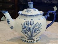 Side view showing embossed tulip design on white ceramic with blue gradient rim, handle, and spout accents.