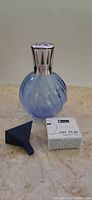 Photo of assembled Lampe Berger lamp with silver metal crown cap on top, showing the blue swirled glass body and black funnel, with fragrance box next to it.