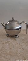 Front left angled view showing the spherical metal teapot with three ball feet, curved handle and spout, and domed lid with round knob