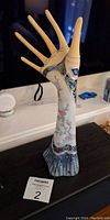Full view of the resin hand jewellery holder showing the arm and fingers spread, decorated with floral accents and blue ornamental patterns at the base.