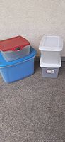 Four storage bins arranged outside against a wall on concrete. Bins are blue with blue lid, clear with red lid, clear with white lid, and gray with white lid. Various sizes.