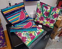 Two used large reusable shopping bags lying flat, one with vibrant blue, yellow, white, and purple stripes and watermelon slice accents, the other with tropical floral pattern in pink, yellow, and green.