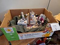 Wide view of all three porcelain figures with piano and harp resting in a cardboard box lined with a green blanket for protection.