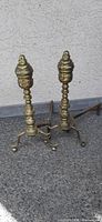 Pair of Victorian era brass andirons standing on textured gray surface against stucco wall background. Both have turned baluster style shafts topped with bee hive shaped finials and three curved legs ending in ball feet. Horizontal iron fire bars extend rearward.