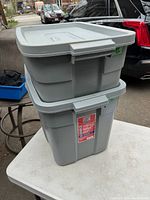 Two stacked gray Rubbermaid Roughneck totes with lids, shown outside on a table, highlighting their size difference and condition.