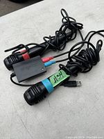 Top view of two black wired SingStar microphones with USB and audio jack adapters and color bands near microphones, all cables coiled, placed on a light surface.