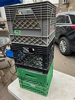 Three stacked plastic milk crates in grey, black, and green on a white table outside, showing open lattice sides and handles.