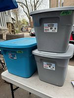 Three plastic storage totes stacked on an outdoor table: 1 large blue tote and 2 smaller gray totes, all with lids and Sterilite labels.