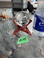 Clear crystal ball displayed on a reddish-brown folding wooden stand placed on a glass table outdoors, with other objects like a water bottle and decorative dog figurine nearby.