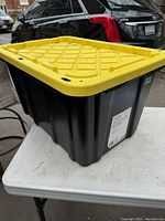 Side view of black HDX tough tote with yellow lid on a white table, showing sturdy plastic construction and reinforced lid design.