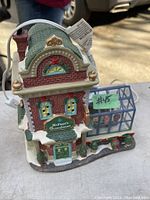 Photo of a hand-painted ceramic Victorian-style McFaye's Greenhouse decorative figurine with light-up feature. Shows red brick building with green roof and glass-panel greenhouse section on the side, snow and holiday greenery accents.