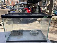 Front view of rectangular glass tank with black frame and ventilation mesh top lid, showing rock and bowl decorations inside.