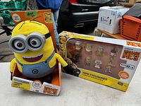 Minion Dave plush toy with overalls and goggles displayed next to a boxed set of mini figurines from Despicable Me 3.