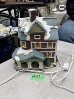Front view of a porcelain Victorian house figurine showing details of roof, chimney, windows with flower boxes, and porch with snow accents. White electrical cord with switch visible to operate light feature.
