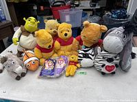 Plush toys arranged on a table including multiple Winnie the Poohs, Shrek, Eeyore, Nemo, a zebra plush, and other stuffed animals.