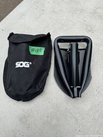 Folded shovel alongside black SOG carrying pouch on white surface.
