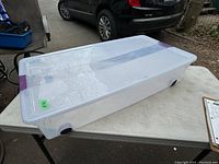 Image showing clear plastic underbed storage tote with translucent lid and purple latches on a table outdoors.