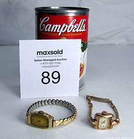 Two antique wristwatches placed side by side, one with a stretch metal band and rectangular yellowed dial, the other a small rectangular watch with a delicate chain band.