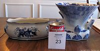 Two white porcelain pieces with blue glaze, one wider oval shallow accent pot with blue floral pattern, one large flared mouth vase with blue 3D lily, frog, and salmon details