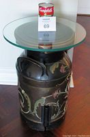 Full front view of black metal milk can painted with floral folk art design and thick round glass top shown with a Campbell's soup can for scale.