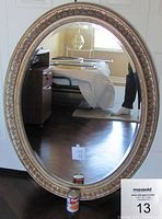 Front view of the large oval bevelled mirror against a white door, showing ornate floral frame design and bevelled glass reflecting room interior.