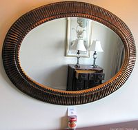 Photo front view of a large oval mirror on wall showing its textured dark brown/bronze frame with ridged pattern and bevelled mirror surface.