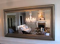 Large rectangular bevelled glass mirror with silver decorative frame hung on a wall reflecting a chandelier and living room furnishings.