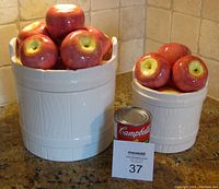 Two cream-colored ceramic canisters designed as apple barrels with red ceramic apple lids. One larger (11"x7.75"), one smaller (7.75"x5.75"). The canisters are displayed on a kitchen counter with a Campbell's soup can for scale and a Maxsold tag.