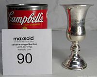Front angle view of the sterling silver cup next to a Campbell's soup can for size reference, showing the polished silver surface and decorative engraving.