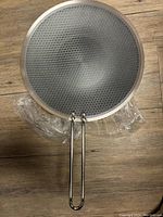 Full view of new 12-inch stainless steel frying pan with honeycomb pattern and double loop handle lying on wooden floor with some plastic wrapping underneath.