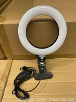 Photo showing the white circular LED ring light mounted on a black heavy-duty clamp with attached USB cable and controller on a brown surface.