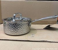 Side view of the hammered textured stainless steel saucepan with lid and long handle on a cardboard surface.