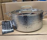 Image of the Cuisinart stainless steel Dutch oven showing hammered exterior surface and fitted lid with handle, sitting on cardboard boxes.