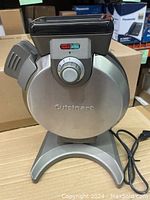 Front view of vertical waffle maker showing brushed stainless steel body, control knob with numbered settings, power cord, and indicator lights.