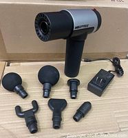 Photo of Maxfree massage gun with six massage heads and charger laid out on cardboard surface showing all components.